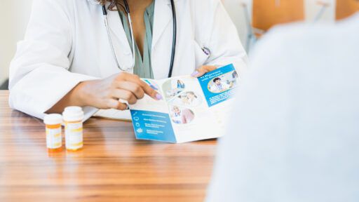 A female doctor shows an unrecognizable patient a brochure, detailing healthcare or pharmaceutical options available to the patient.
